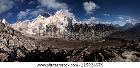 Panoramic view of Mt Everest from Kala Patthar in dry landscape with snow capped himalayas and blue sky, and rocky foreground.