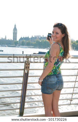 Smiling woman looking at camera holding mobile phone, London attractions in background