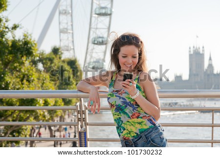 Woman looking at phone smiling, texting from bridge over River Thames, London with attractions in background
