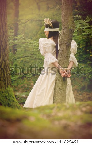 Dark fairytale inspired image, beautiful young woman in woods, tied with rope to a tree and blindfolded