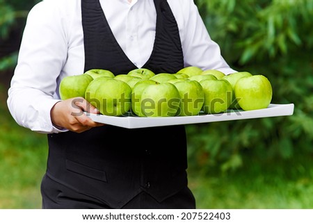 Green apples on tray. Waiter holding a tray with green apples. Shot outdoor in the garden on a green background.