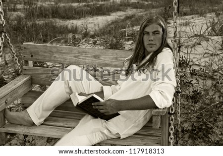 Young male caucasian, 21 years old, sitting on bench swing reading a book. Photographed in the dunes of Tybee Island, Georgia, USA.