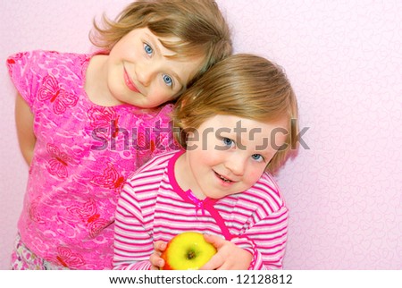 Two sister standing next to each other, face to face. They both dressed in pink clothes and they happily smile. In the hands of the child\'s great apple. Siblings.