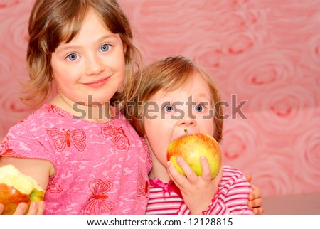 Two sister standing next to each other, face to face. They both dressed in pink clothes and they happily smile. In the hands of the child\'s great apple.