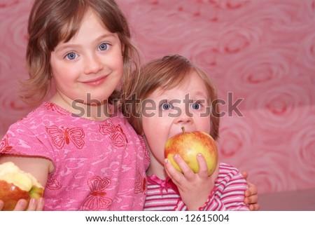 Two sister standing next to each other, face to face. They both dressed in pink clothes and they happily smile. In the hands of the child\'s great apple.