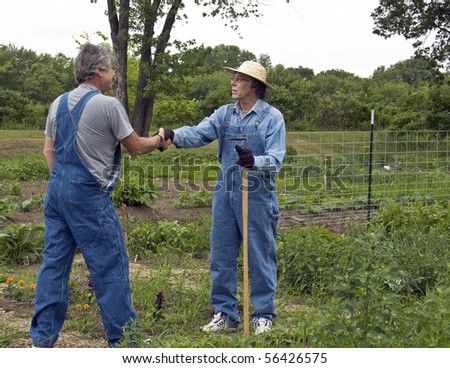 two men in bib overalls greet each other in a garden with a handshake