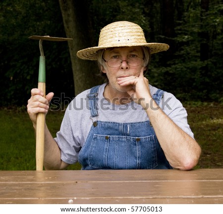 farmer in straw hat and bib overalls holding a hoe sitting at a table