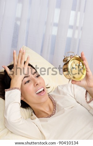 Shocked beautiful young woman  looking at an alarm clock  while lying in the bed.