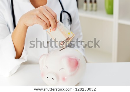 Closeup of female doctor hand putting a fifty Euro in the Piggy Bank.