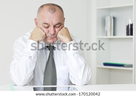Worried businessman with hand on his chin sitting at the desk and looking down.