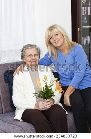 Smiling mature nurse sitting on the sofa and embracing senior woman ,holding a bouquet of flowers.