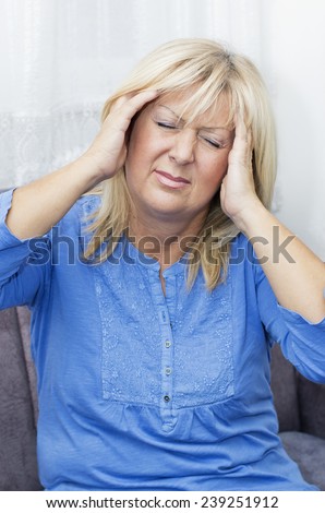 Mature blond haired woman with eyes closed sitting on the sofa  and holding  her  head in pain.