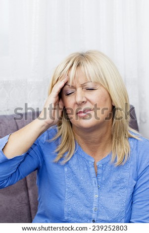 Mature blond haired woman with eyes closed sitting on the sofa  and touching her  head in pain.