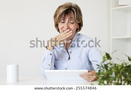 Surprised  mature businesswoman with hand covered her mouth looking at digital tablet while sitting at desk in the office.
