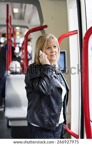 Mature Blond haired woman  using a  public transport on his way to work.  She standing next to door and using a smart phone. People in the background.