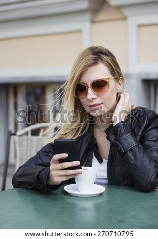 Blond haired young  woman with sunglasses sitting in the street cafe drinking a coffee and using a smart phone.