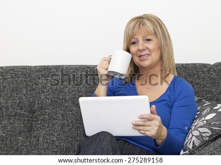 Charming mature woman sitting on the sofa in the living room drinking a coffee and surfing the net.