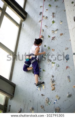 A girl wearing harness and belaying rope and climbing on a very high rock climbing wall.