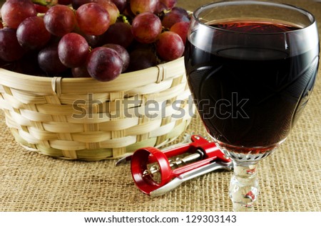 Red grapes in a basket wiht a glass of red wine and corkscrew on a jute texture