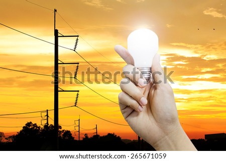 Hand holding a glowing light bulb and electricity pole sunset background, power energy