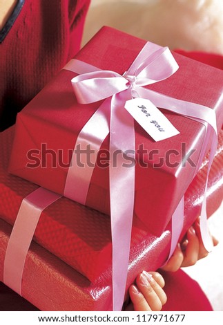 stacks of gifts wrapped in red with a pink ribbon around the whole, held by a woman dressed in red
