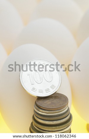 a south korean hundred won coin standing at the top of stack of coins leaning against white egg