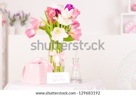 Picture of fresh tulip flowers in beautiful vase with paper greeting postcard on the table, little pink gift box with ribbon, home interior, happy mothers day, festive still life, love concept