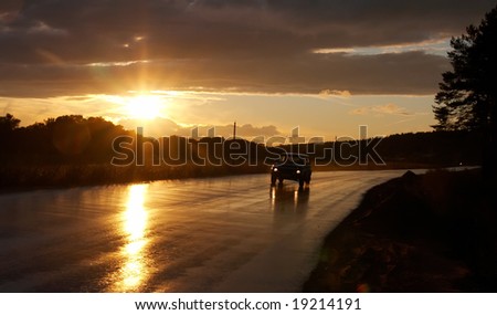 Lone car driving on a wet road against setting sun and a cloudy sky