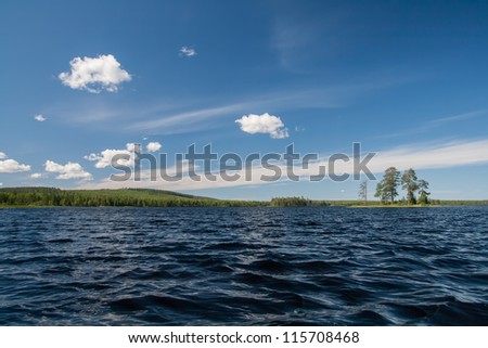 Landscape of a lake in northern Russia. Dark waters, bright blue sky, rolling hills on the distant shore and a lone island for a few trees.