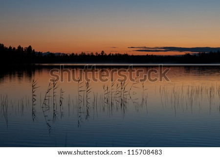 Quiet still waters of a lake, with some grass growing out of water and reddened sky in background