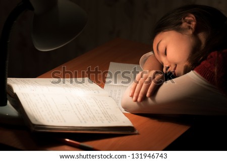 Girl asleep at a table doing homework by the light of the lamp