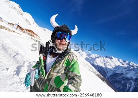 Bearded skier in a green outfit with horns with a can in his hands on top of snow-capped mountains against the blue sky