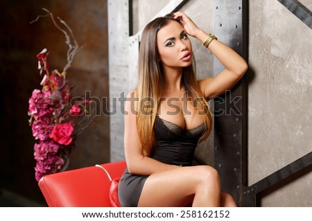 Beautiful young girl with big breasts, full lips, straight hair in a short black dress, athletic, tanned, sitting on a red leather couch in the studio on a background of a wall decorated with metal