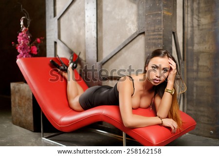 Beautiful young girl with big breasts, full lips, straight hair in a very short black dress, athletic, tanned, lying on a red leather couch in the studio on a background of a wall decorated with metal