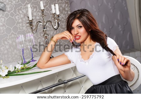 Beautiful leggy brunette girl with big breasts, full lips in a short black dress with a white collar and cuffs in high heels standing near bouquets of roses in vases in white interior