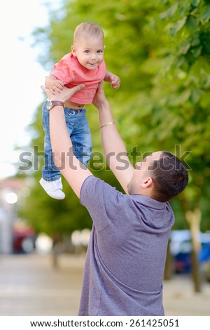 1 year baby in the arms of his father, blond, gray eyes, a red shirt, blue jeans. Against the background of green trees. A young father playing with his son.