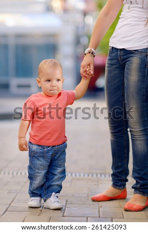 1 year baby, holding the hand of his mother, blond hair, gray eyes, a red shirt, blue jeans, mother in a white T-shirt, blue jeans. Against the background of green trees.