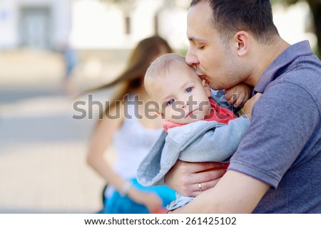 Young father kissing his son. 1 year baby in the arms of his father, blond, gray eyes, a red shirt, blue jeans. Against the background of the cityscape.
