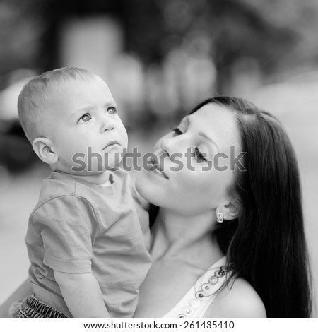 Black and white, 1 year baby in the arms of mother, blond, gray eyes, jeans, Mum young beautiful brunette with straight hair, a white T-shirt. Young mother with her baby in her arms.