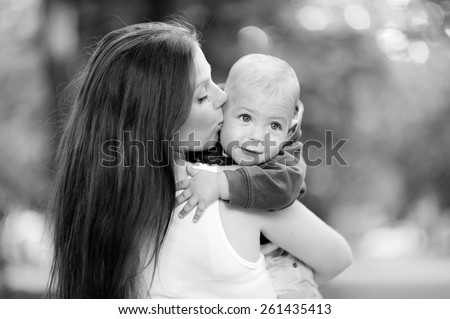 Black and white, 1 year baby in the arms of mother, blond, in a jacket, jeans, Mum beautiful brunette with straight hair, a white T-shirt. Young mother with her baby in her arms.