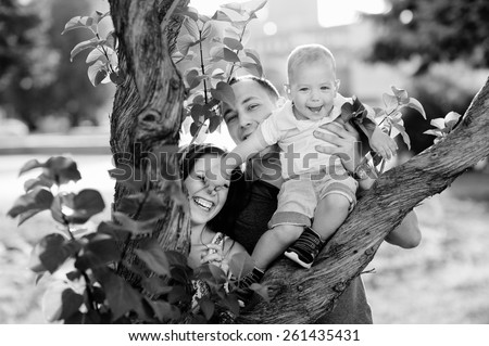 Black and white, happy family: mother, father, son, 1 year old, in a jeans, white shirts, smiling, happy, love. Next to a tree in a city park.