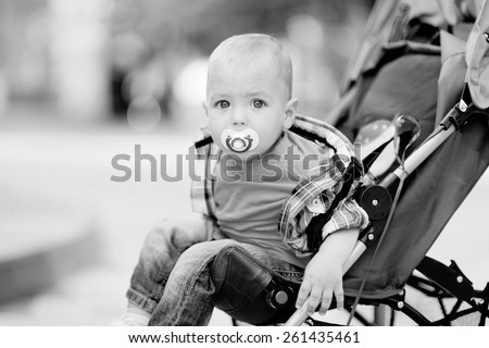 Black and white, 1 year baby sitting in the baby carriage, with a pacifier in his mouth, blond hair, plaid shirt, jeans, a city on the background of trees. Baby with a pacifier.