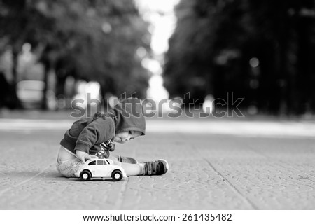 Black and white, 1 year baby sitting on the sidewalk in a jacket with a hood with white print in shorts and sneakers, plays a toy car on a background of trees and the city.