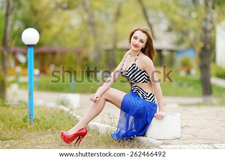 beautiful sexy brunette girl in short striped black and white dress with a blue plume, red high heel shoes, with big Breasts and long legs on the street in the Park. Spring walk. Summer walk.