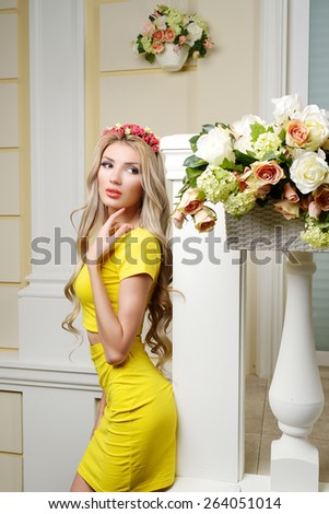 Beautiful slim sexy girl with big lips, big breasts and long curly hair in yellow Sexy Costumes with flowers in her hair near the terrace decorated with flowers.  Lady in the interior. Spring woman