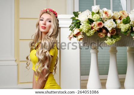 Beautiful slim sexy girl with big lips, big breasts and long curly hair in yellow Sexy Costumes with flowers in her hair near the terrace decorated with flowers.  Lady in the interior. Spring woman
