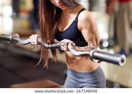 Brutal athletic woman pumping up muscles with barbell. Attractive fitness woman, trained female body. Brunette sexy fitness girl in black and gray sport wear with perfect body in the gym.