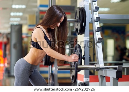 Athletic girl doing exercises with barbell in gym. Attractive fitness woman, trained female body. Brunette sexy fitness girl in black and gray sport wear with perfect body in the gym.