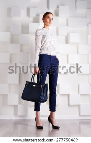 stylish fashionable girl with smoothed hair wearing blue breeches and shirt with print in high heels with a blue bag on the background of white squares in the Studio