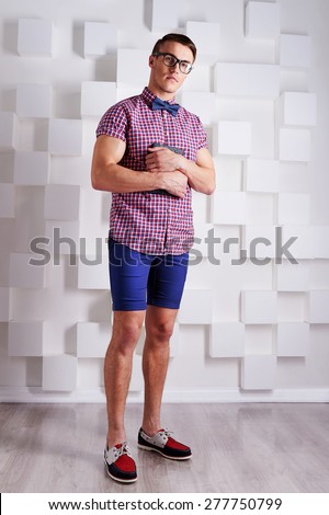 stylish trendy athletic guy in tight blue shorts and a plaid shirt with bow tie in points in the multi-colored loafers with the book with the white squares in the Studio